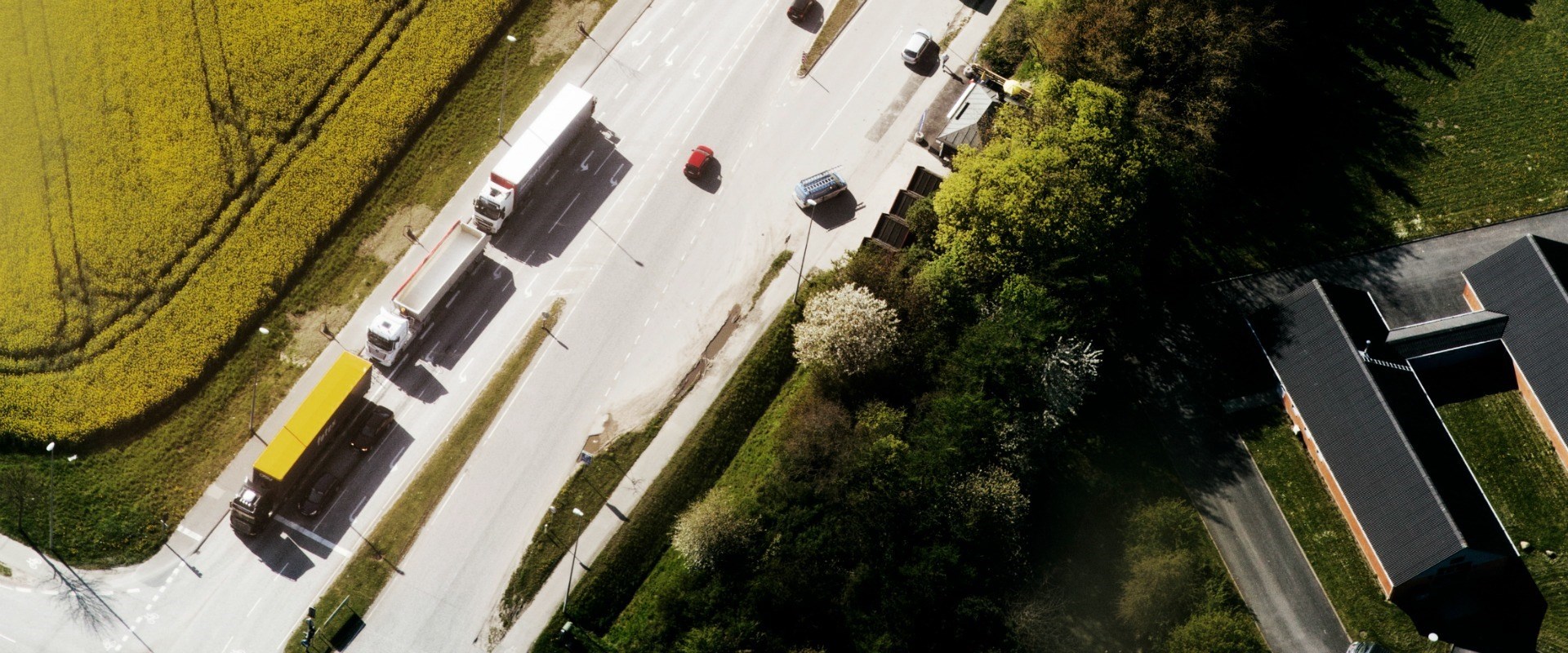 Luftfoto af en vej med tre lastbiler og fire personbiler. På begge siden af vejen er der grønne områder.